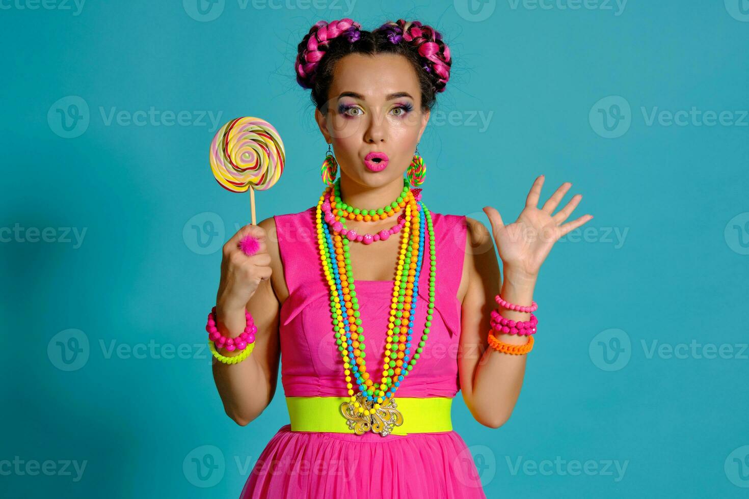 encantador niña con un multicolor trenzas peinado y brillante constituir, posando en estudio en contra un azul fondo, participación un pirulí en su mano. foto