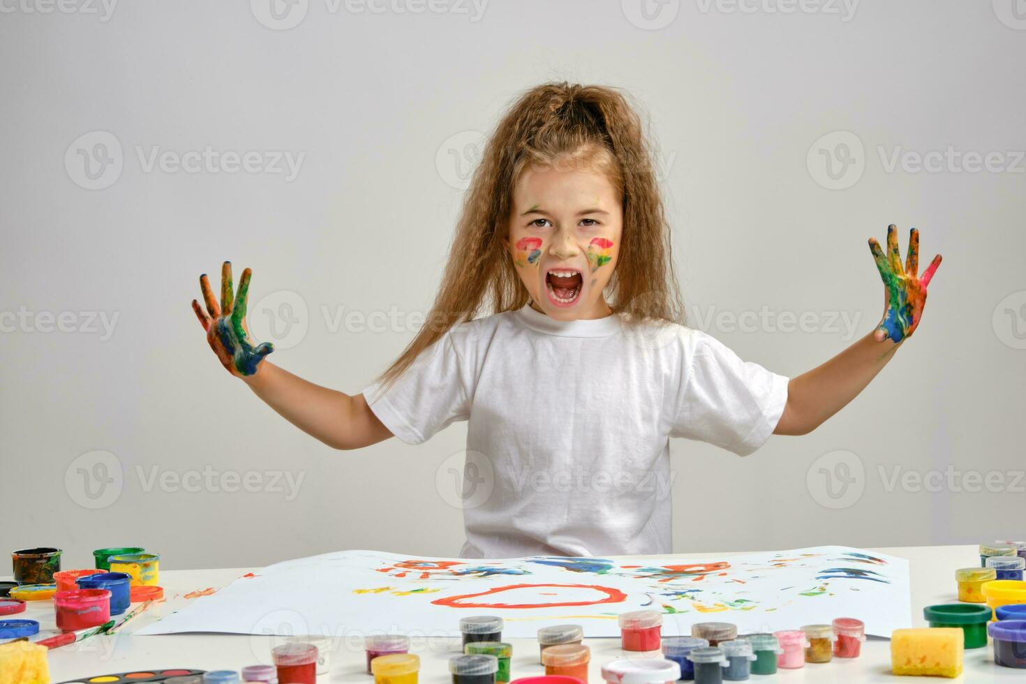 Little girl in white t-shirt sitting at table with whatman and paints on it, posing with painted face and hands. Isolated on white. Medium close-up. photo