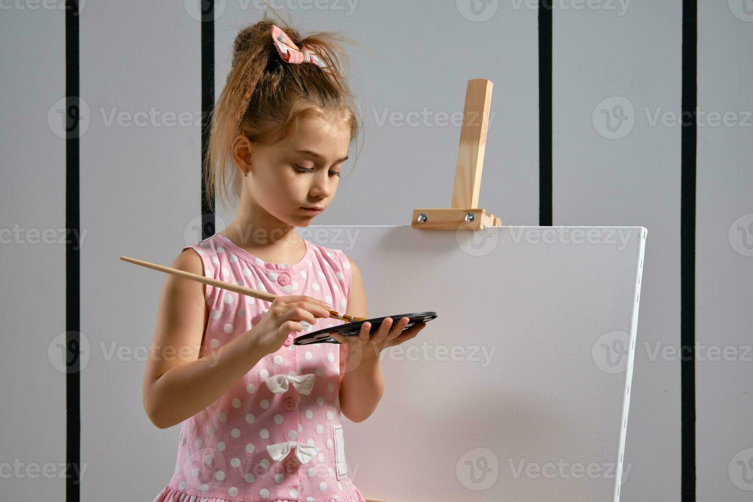 Little girl artist in a pink dress is standing behind easel and painting with brush on canvas at art studio with white walls. Medium close-up shot. photo