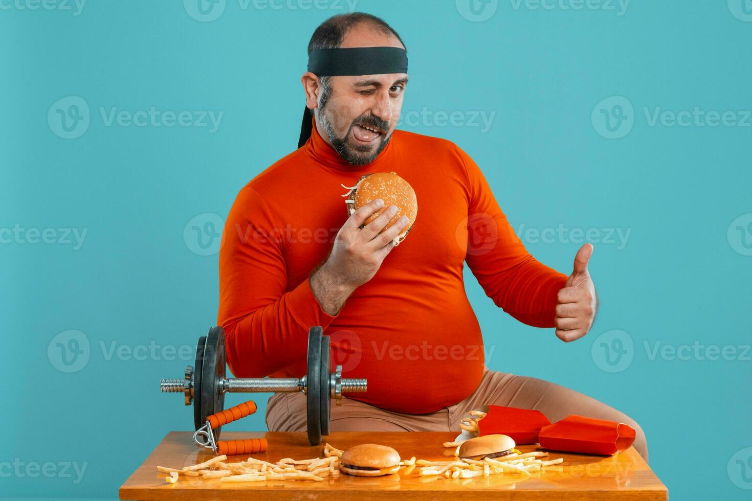 Middle-aged man with beard, dressed in a red turtleneck, headband, posing with burgers and french fries. Blue background. Close-up. Fast food. photo