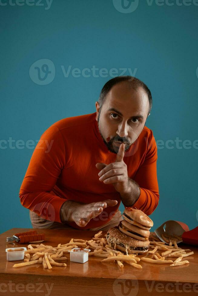 Close-up portrait of a middle-aged man with beard, dressed in a red turtleneck, posing with burgers and french fries. Blue background. Fast food. photo