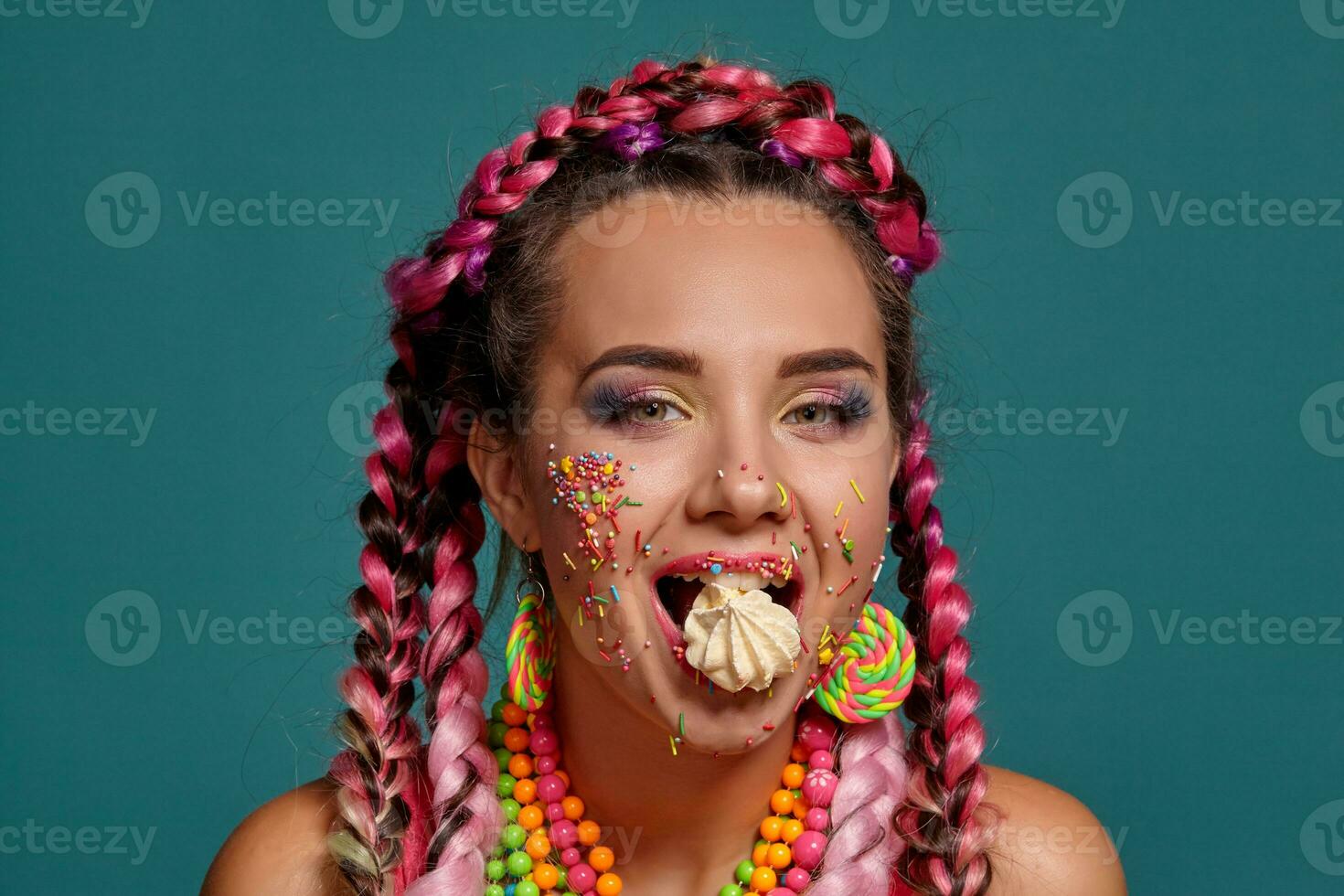 Lovely girl with a multi-colored braids hairstyle and bright make-up, posing in studio against a blue background, holding a marshmallow in her mouth. photo