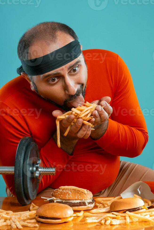 Middle-aged man with beard, dressed in a red turtleneck, headband, posing with burgers and french fries. Blue background. Close-up. Fast food. photo