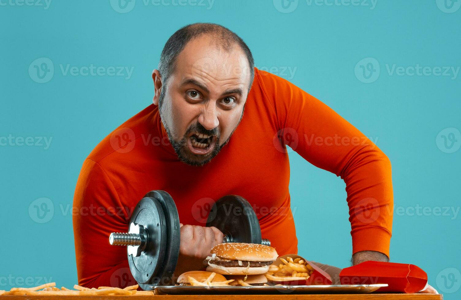 Close-up portrait of a middle-aged man with beard, dressed in a red turtleneck, posing with burgers and french fries. Blue background. Fast food. photo