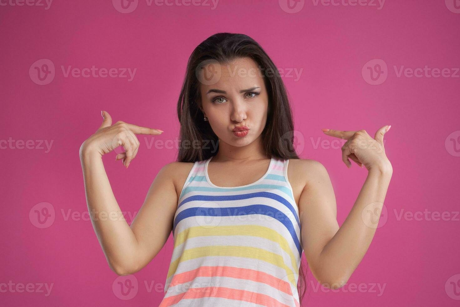 morena mujer con largo cabello, vestido en vistoso a rayas camisa, posando en contra rosado estudio antecedentes. sincero emociones de cerca. foto