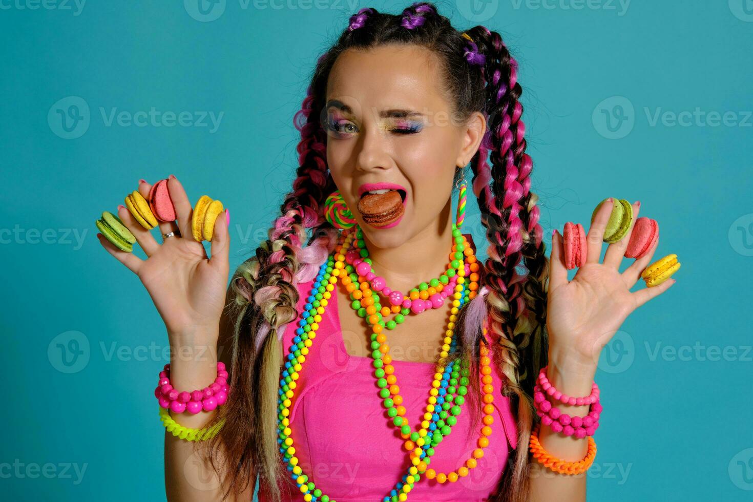 Lovely girl with multi-colored braids hairstyle and bright make-up, holding macarons between her fingers, posing in studio against a blue background. photo