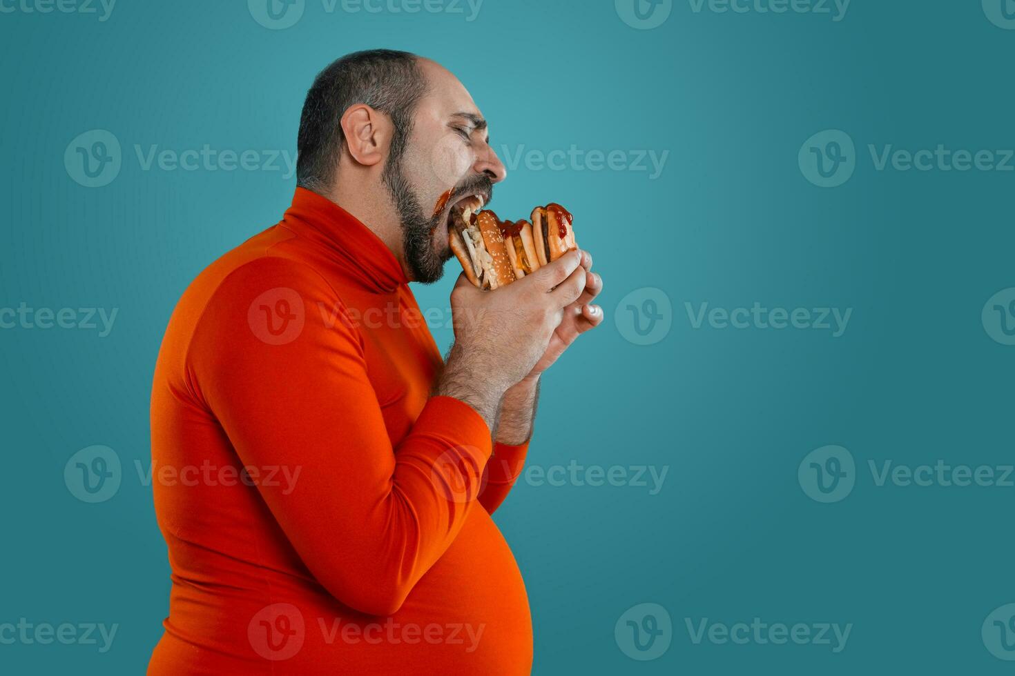 Close-up portrait of a middle-aged man with beard, dressed in a red turtleneck, posing with burgers against a blue background. Fast food. photo