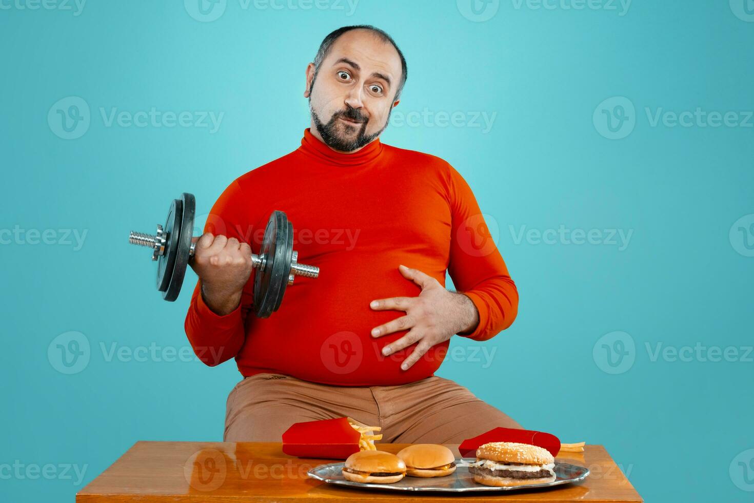 Close-up portrait of a middle-aged man with beard, dressed in a red turtleneck, posing with burgers and french fries. Blue background. Fast food. photo