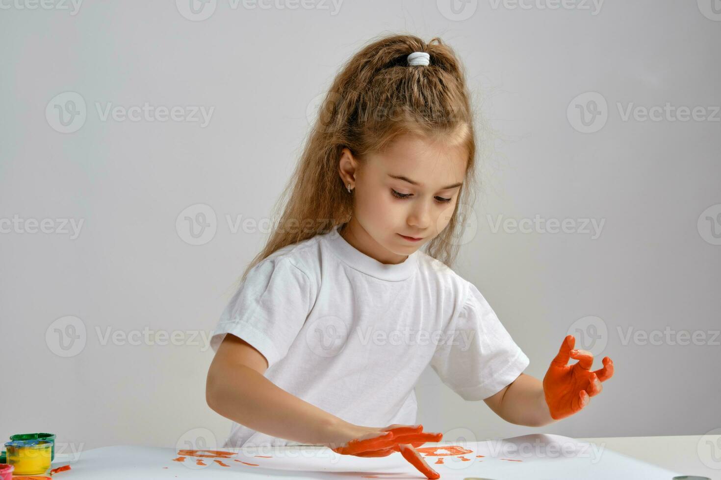 Little girl in white t-shirt sitting at table with whatman and colorful paints, painting on it with her hands. Isolated on white. Medium close-up. photo