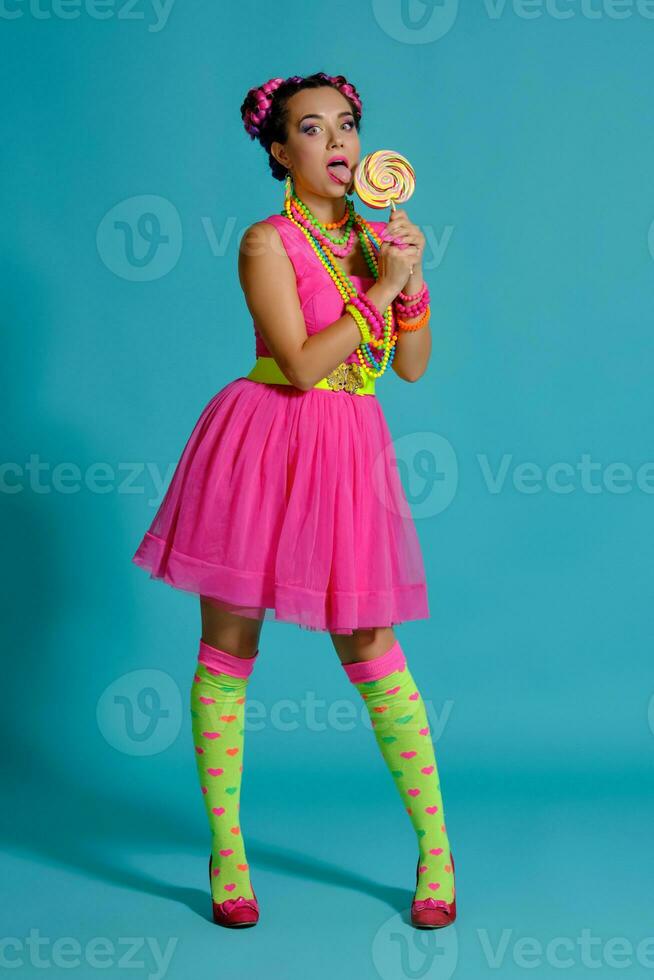 Lovely girl with a multi-colored braids hairstyle and bright make-up, posing in studio against a blue background, holding a lollipop in her hand. photo