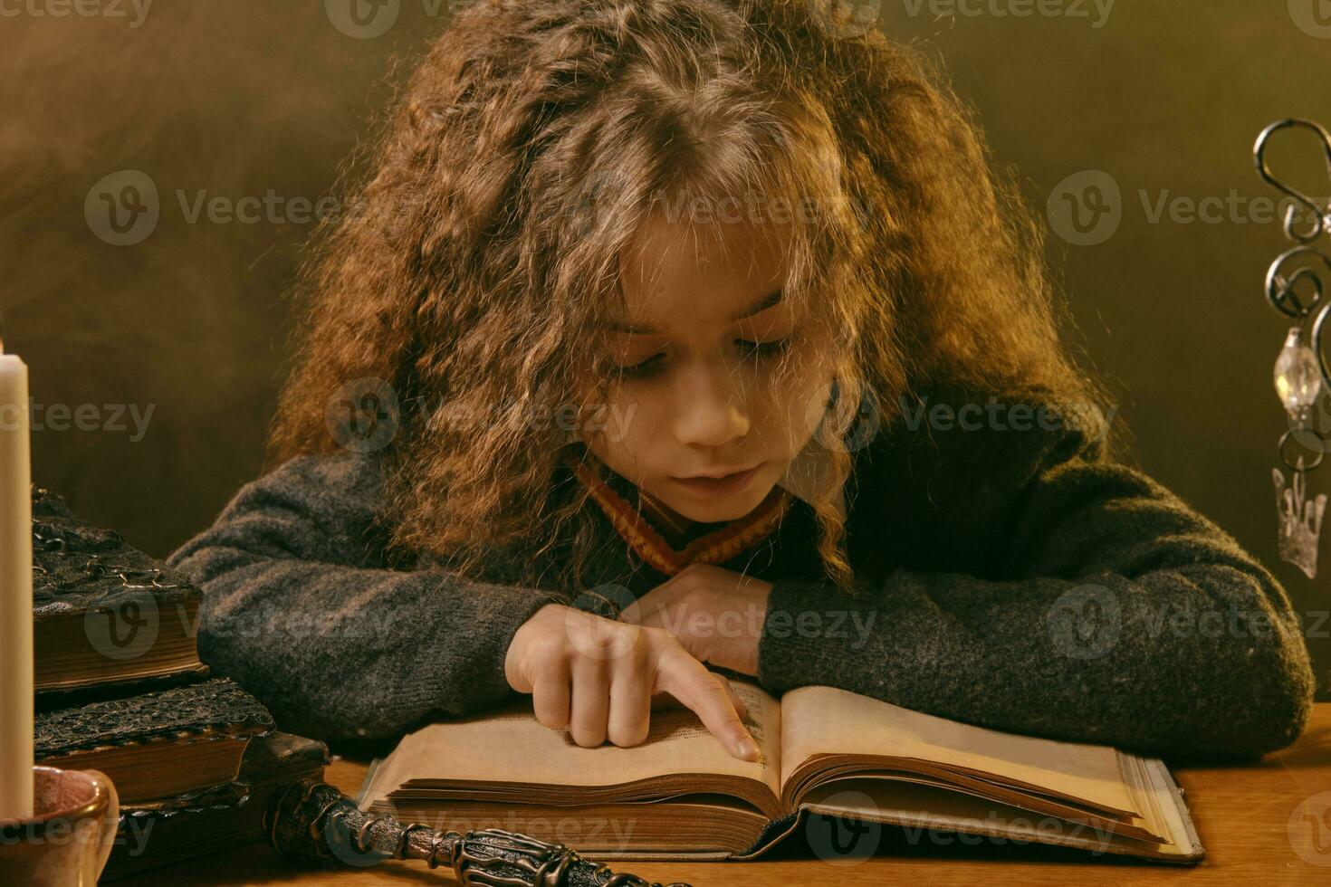 Little witch dressed in dark clothes sitting at the table against black smoky background and reading a book. Close-up portrait. photo
