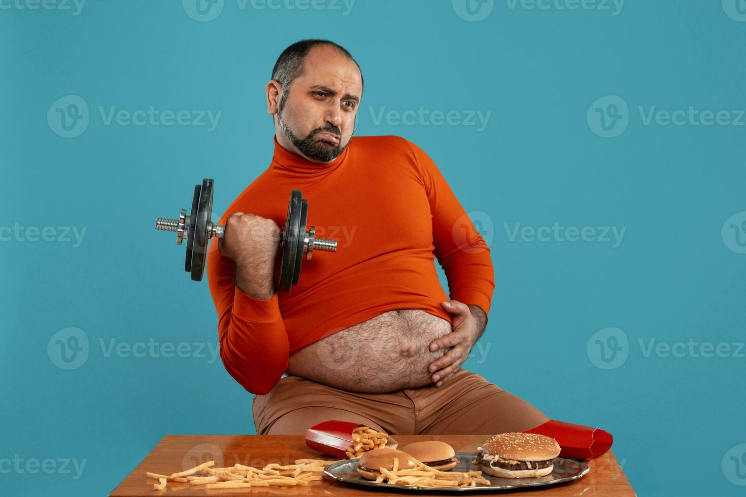 Close-up portrait of a middle-aged man with beard, dressed in a red turtleneck, posing with burgers and french fries. Blue background. Fast food. photo