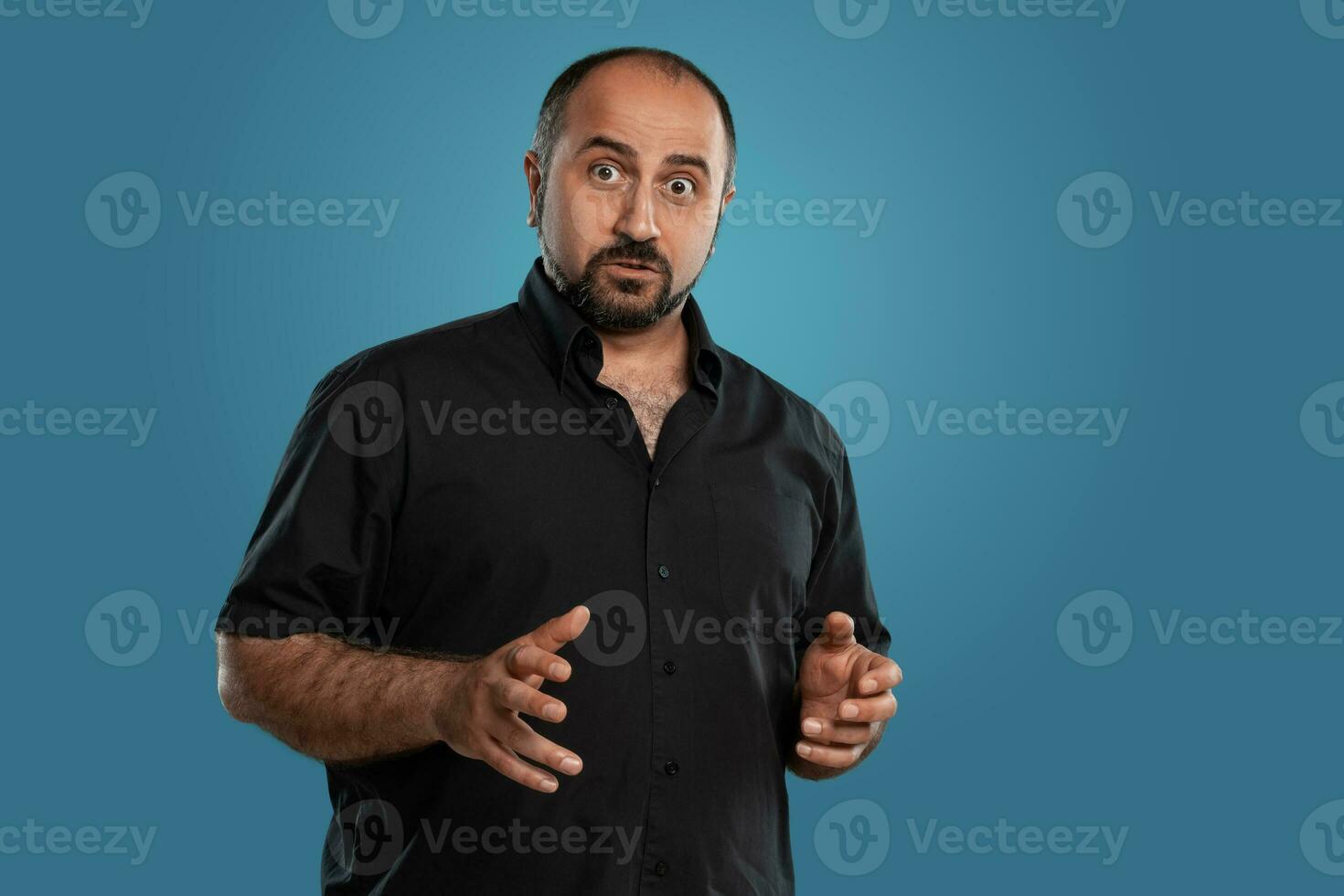 Close-up portrait of a brunet middle-aged man with beard, dressed in a black t-shirt and posing against a blue background. photo