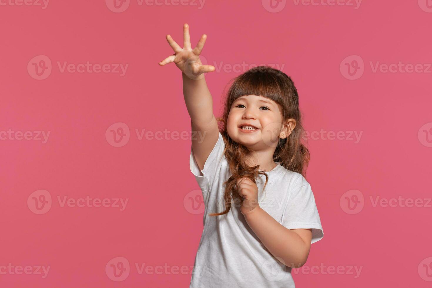 de cerca retrato de un pequeño morena niña vestido en un blanco camiseta posando en contra un rosado estudio antecedentes. sincero emociones concepto. foto