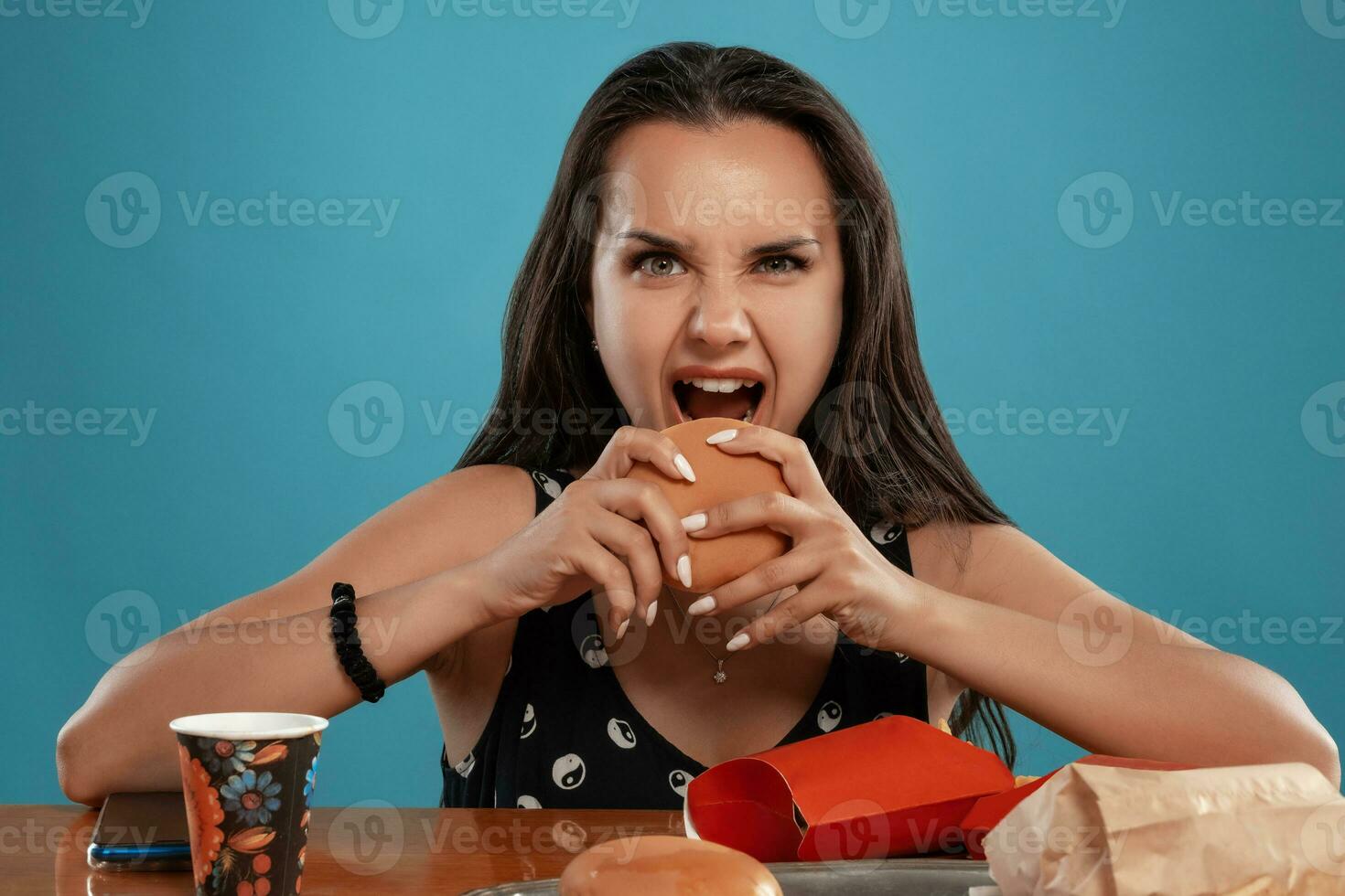 de cerca retrato de un mujer en un negro vestir posando sentado a el mesa con hamburguesas, francés papas fritas y beber. azul antecedentes. rápido alimento. foto