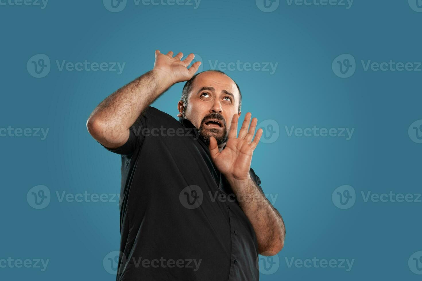 Close-up portrait of a brunet middle-aged man with beard, dressed in a black t-shirt and posing against a blue background. photo