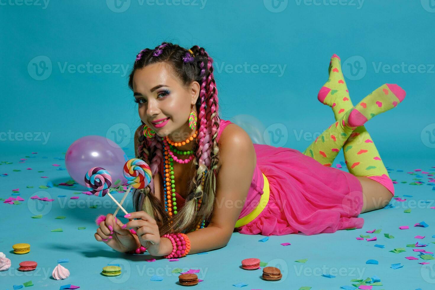 Lovely girl with a multi-colored braids hairstyle and bright make-up, posing in studio with lollipop, air balloons and confetti against a blue background. photo