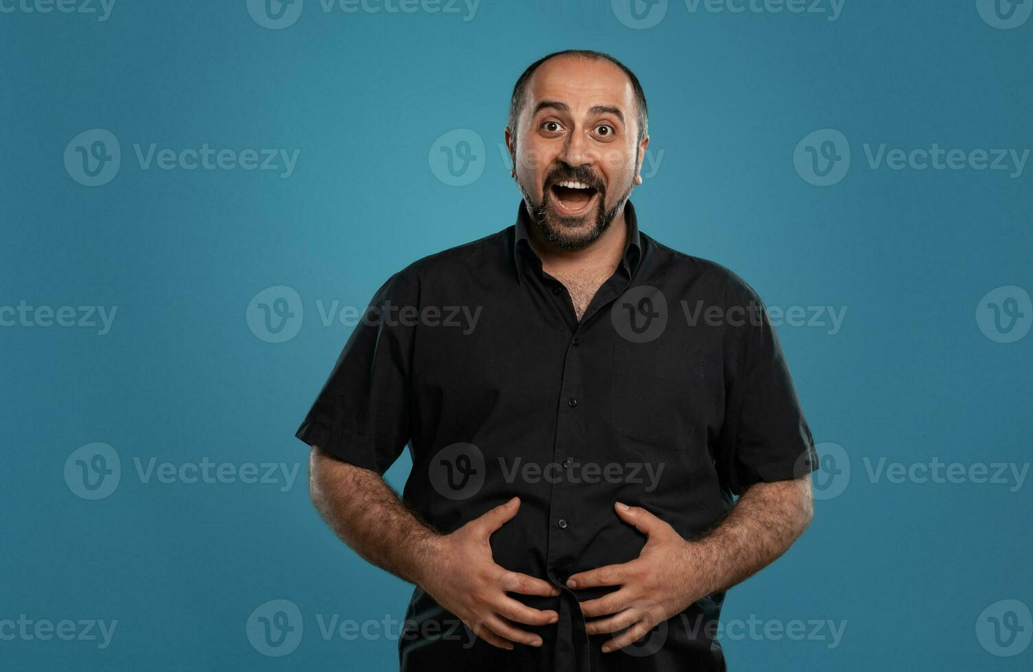 Close-up portrait of a brunet middle-aged man with beard, dressed in a black t-shirt and posing against a blue background. photo