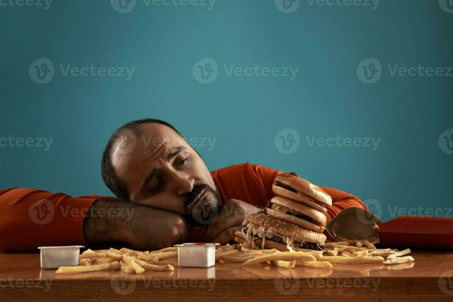 Close-up portrait of a middle-aged man with beard, dressed in a red turtleneck, posing with burgers and french fries. Blue background. Fast food. photo