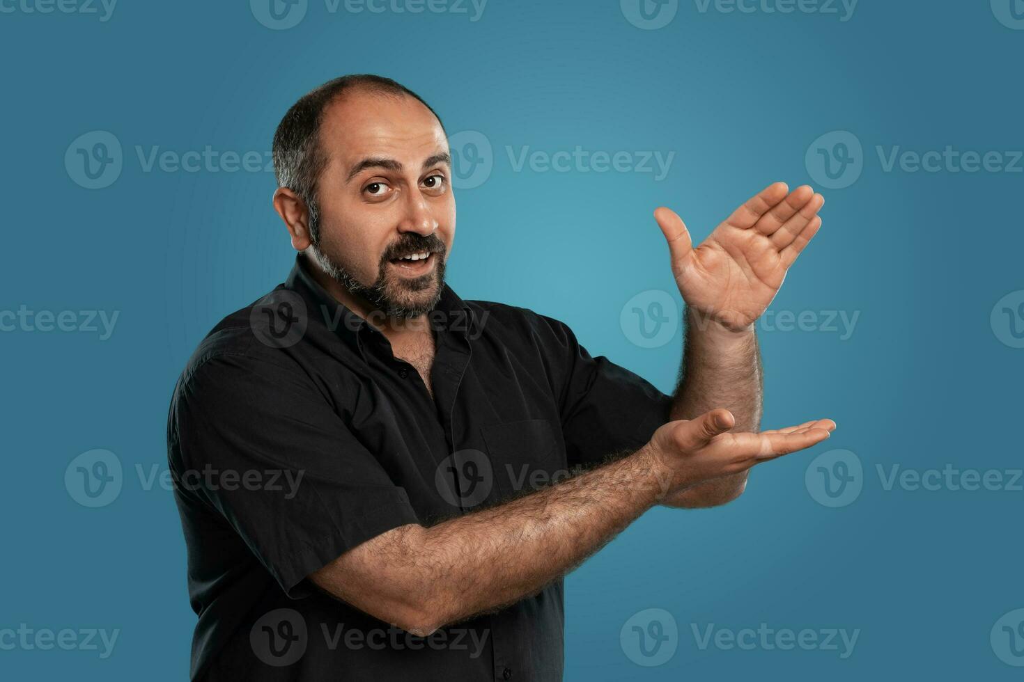 Close-up portrait of a brunet middle-aged man with beard, dressed in a black t-shirt and posing against a blue background. photo