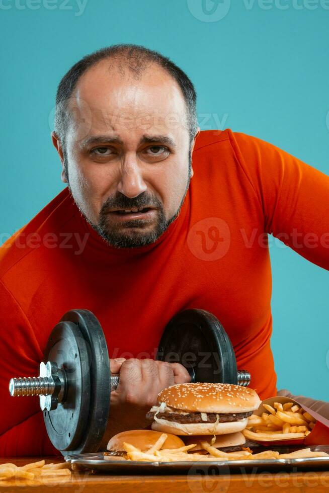 Close-up portrait of a middle-aged man with beard, dressed in a red turtleneck, posing with burgers and french fries. Blue background. Fast food. photo