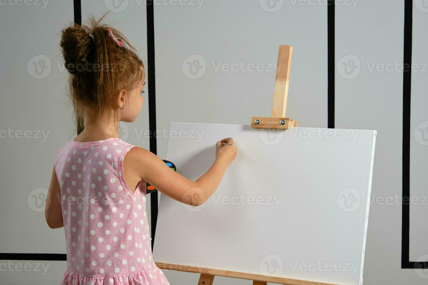 Little girl artist in a pink dress is standing behind easel and painting with brush on canvas at art studio with white walls. Medium close-up shot. photo