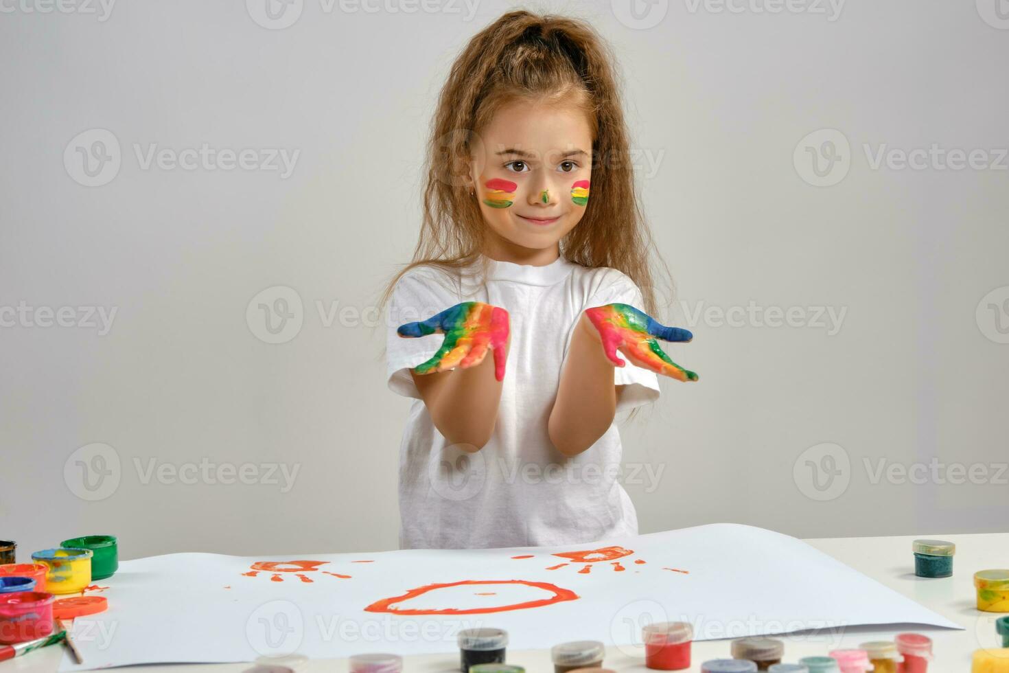 Little girl in white t-shirt sitting at table with whatman and colorful paints, showing her painted hands, face. Isolated on white. Medium close-up. photo