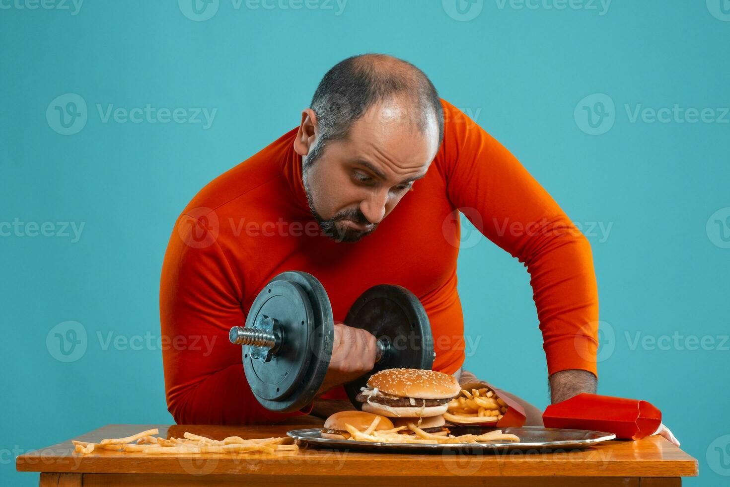 Close-up portrait of a middle-aged man with beard, dressed in a red turtleneck, posing with burgers and french fries. Blue background. Fast food. photo