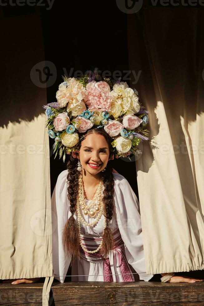 Brunette girl in a white ukrainian authentic national costume and a wreath of flowers is is looking out of the window. Close-up. photo