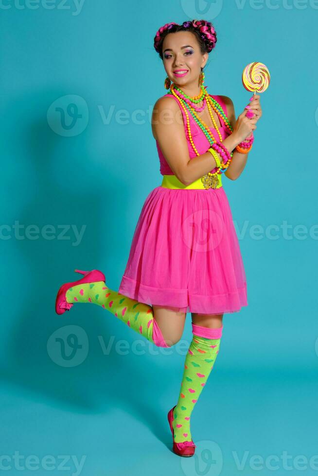 Lovely girl with a multi-colored braids hairstyle and bright make-up, posing in studio against a blue background, holding a lollipop in her hand. photo