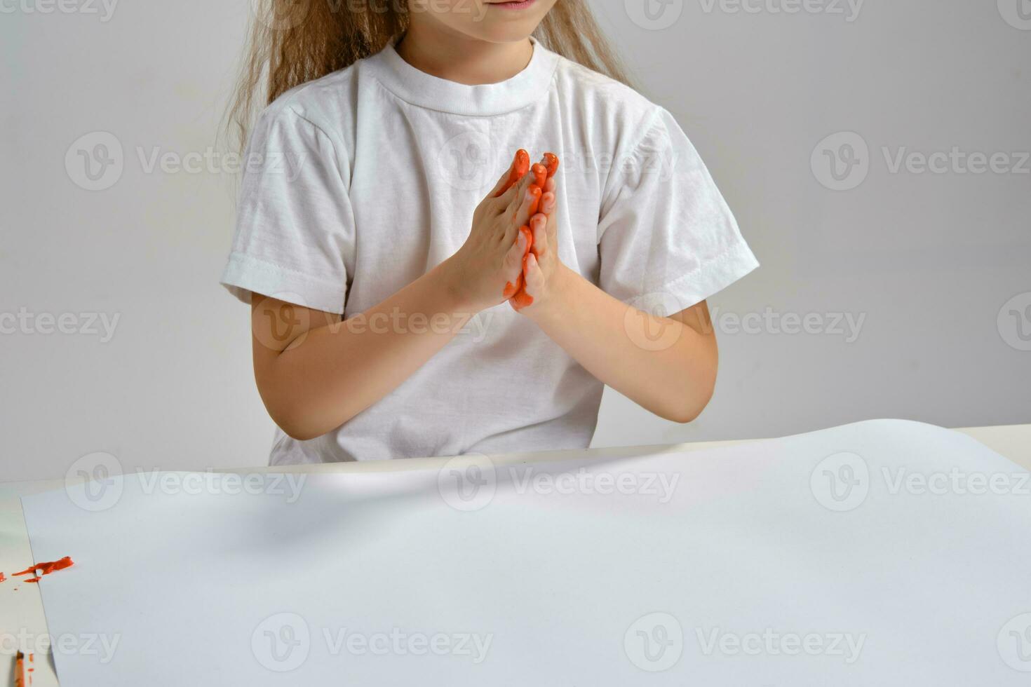 Little artist in white t-shirt standing at table with whatman and colorful paints, painting her hands. Isolated on white. Close-up. photo