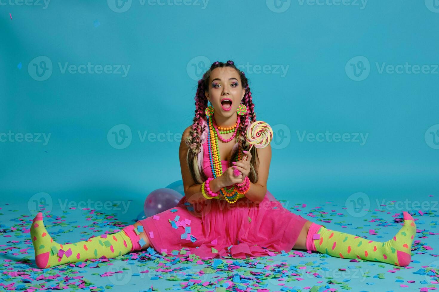 Lovely girl with a multi-colored braids hairstyle and bright make-up, posing in studio with lollipop, air balloons and confetti against a blue background. photo