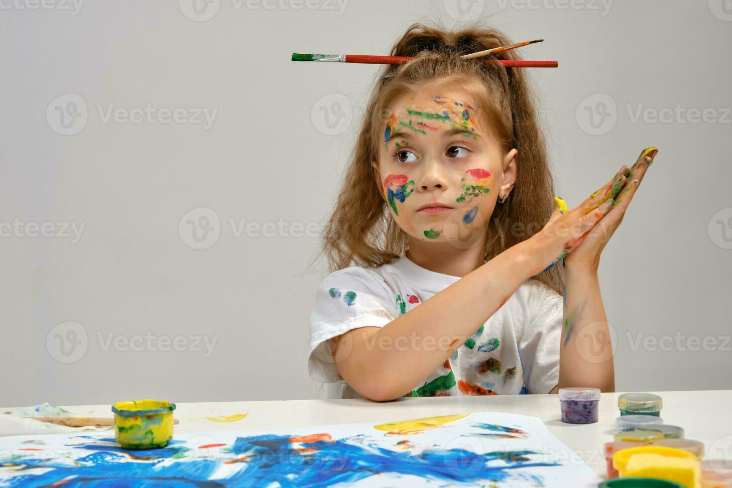 Little girl in white t-shirt, with brushes in her hair is sitting at the table with whatman and paints, painting on it. Isolated on white. Close-up. photo