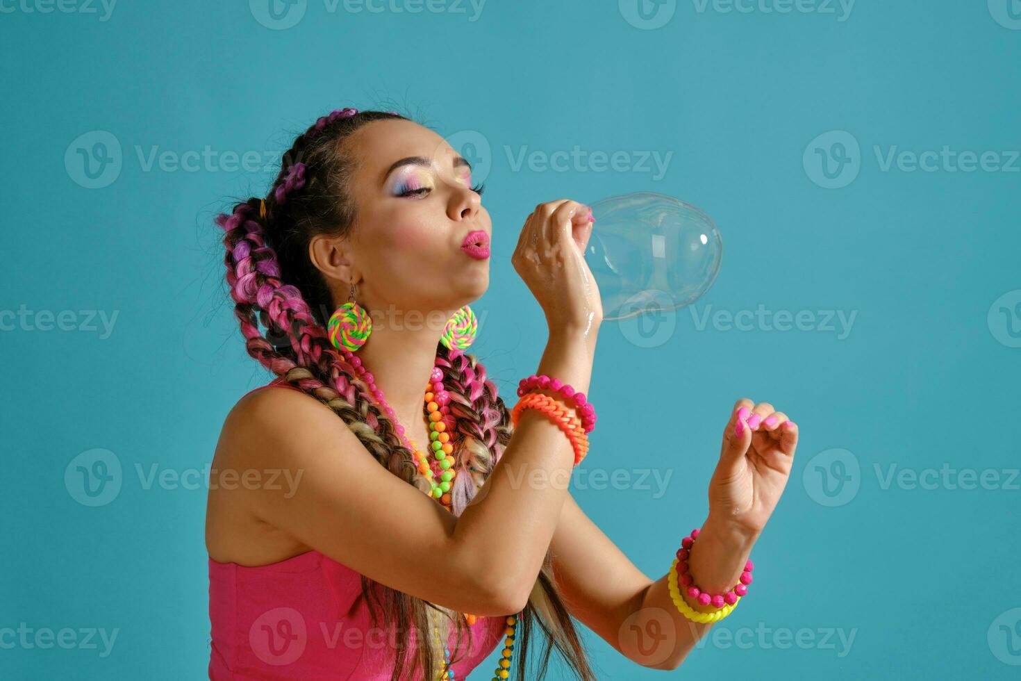 encantador niña con un multicolor trenzas peinado y brillante constituir, es soplo burbujas utilizando su manos, posando en estudio en contra un azul antecedentes. foto