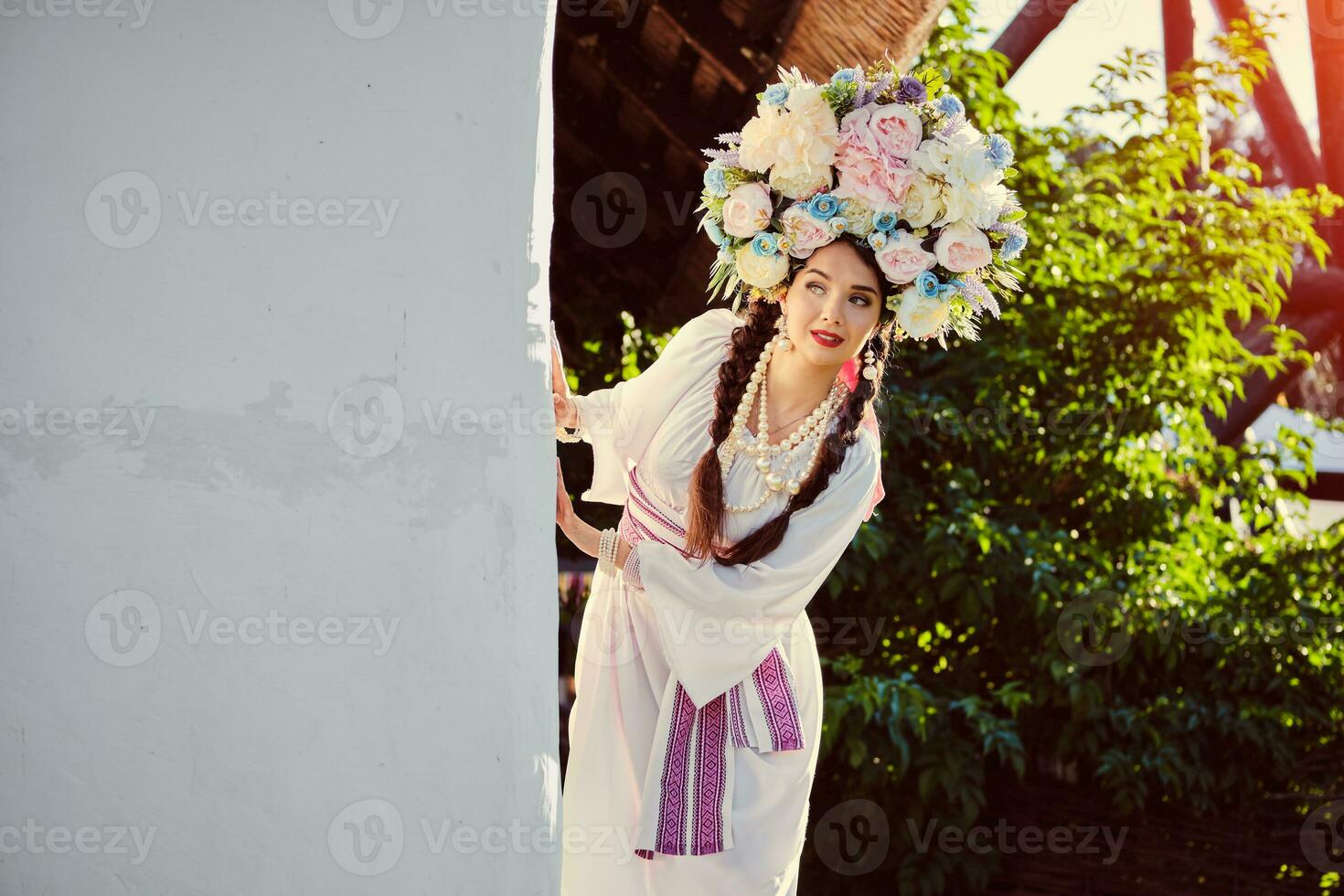 Brunette girl in a white ukrainian authentic national costume and a wreath of flowers is posing against a white hut. photo