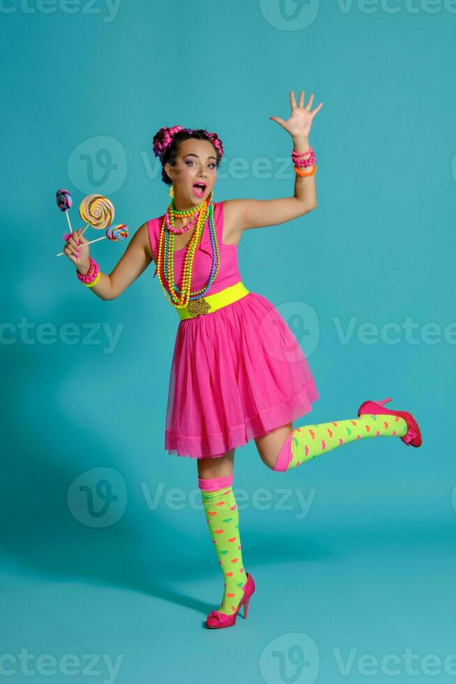 Lovely girl with a multi-colored braids hairstyle and bright make-up, posing in studio against a blue background, holding a lollipop in her hand. photo