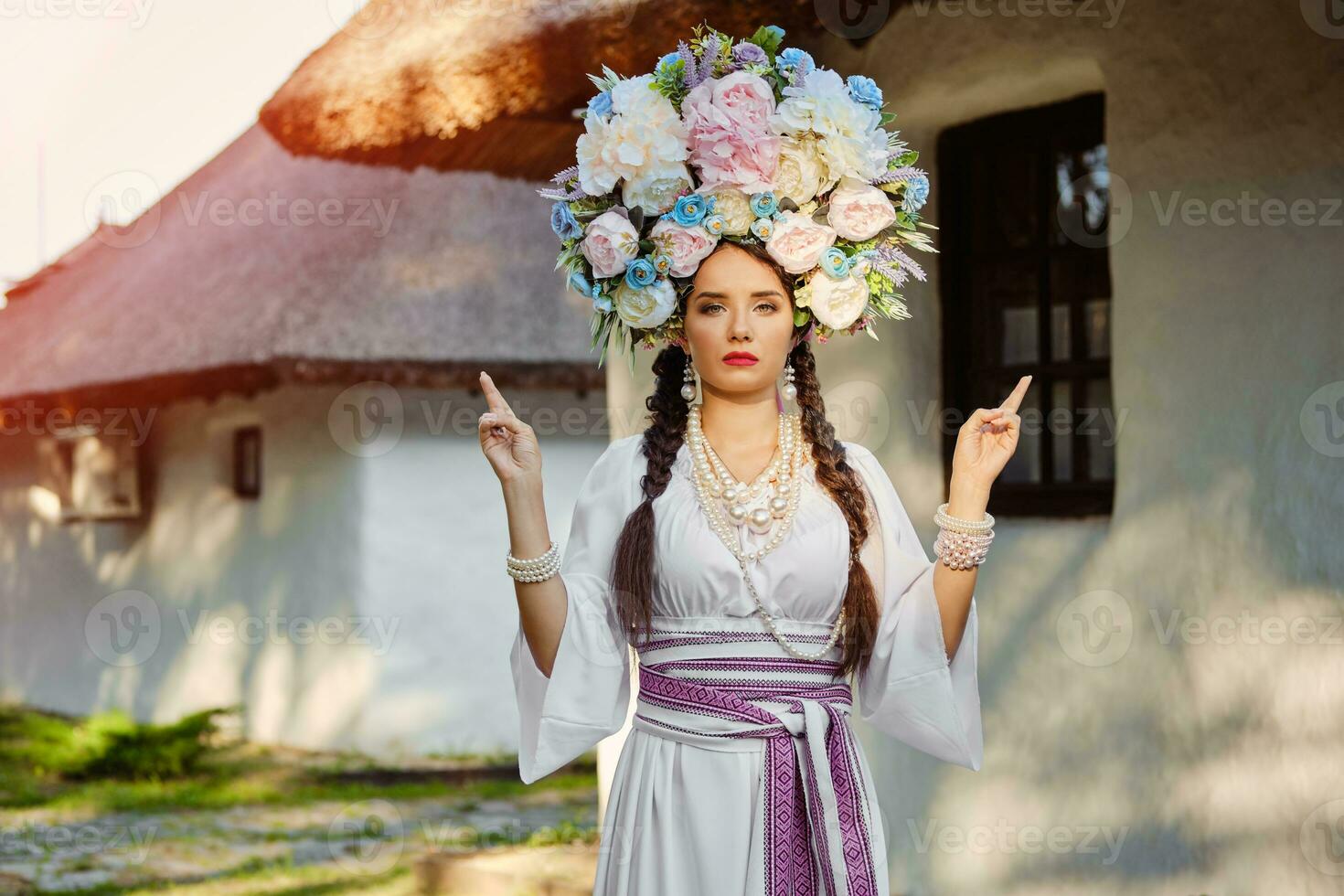 Brunette girl in a white ukrainian authentic national costume and a wreath of flowers is posing against a white hut. Close-up. photo