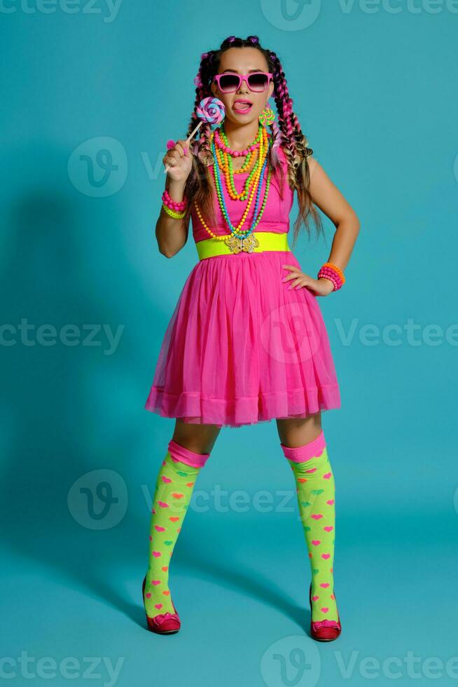 encantador niña con un multicolor trenzas peinado y brillante constituir, posando en estudio en contra un azul fondo, participación un pirulí en su mano. foto