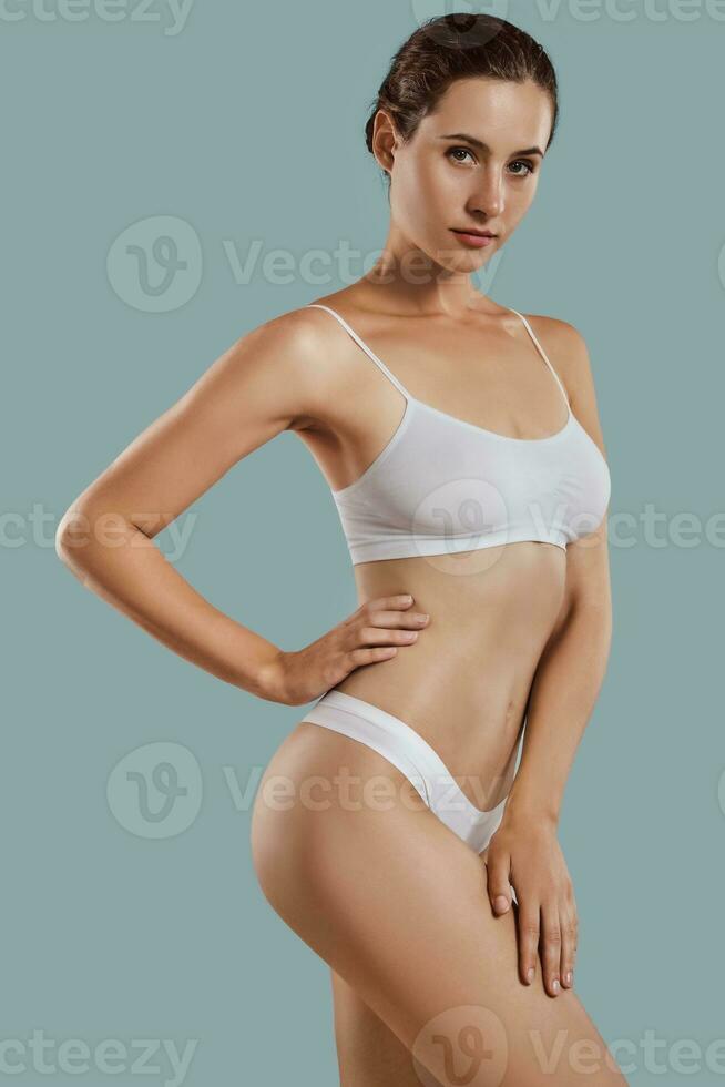 Young woman in white underwear, with bundled hair, hands on hips, posing against gray background. Plastic surgery, aesthetic cosmetology. Close-up. photo