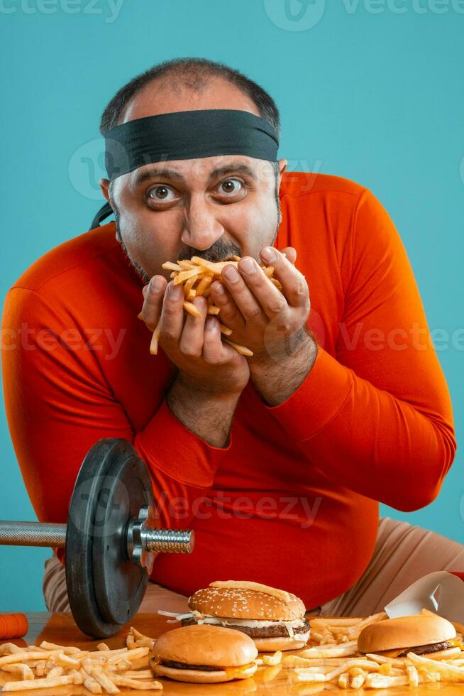 Middle-aged man with beard, dressed in a red turtleneck, headband, posing with burgers and french fries. Blue background. Close-up. Fast food. photo