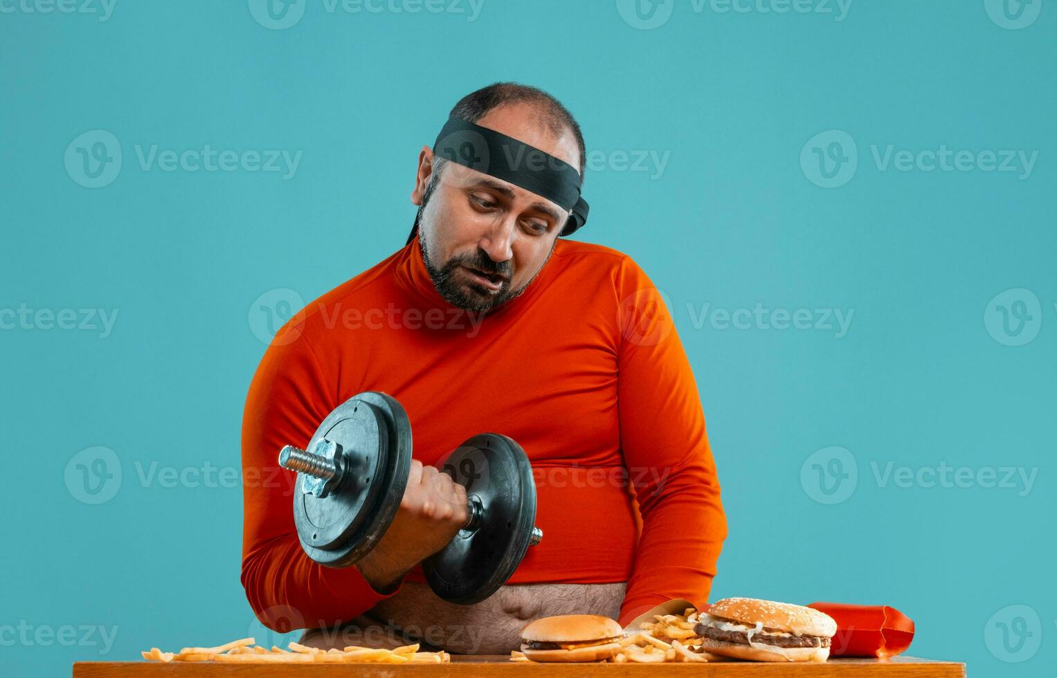 Middle-aged man with beard, dressed in a red turtleneck, headband, posing with burgers and french fries. Blue background. Close-up. Fast food. photo