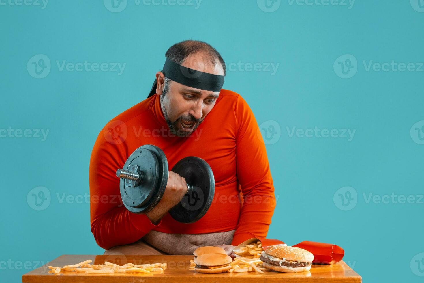 Middle-aged man with beard, dressed in a red turtleneck, headband, posing with burgers and french fries. Blue background. Close-up. Fast food. photo