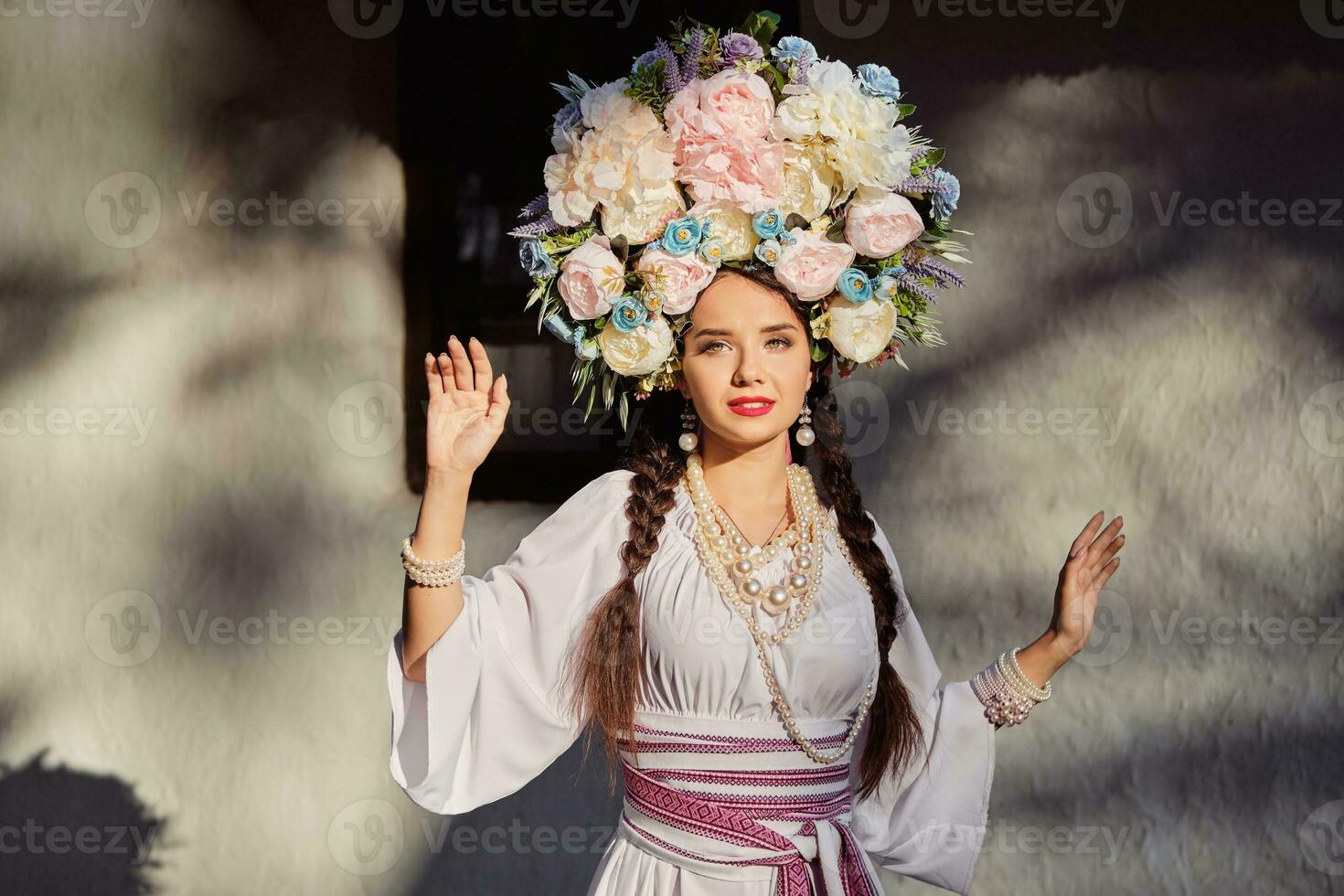 morena niña en un blanco ucranio auténtico nacional disfraz y un guirnalda de flores es posando en contra un blanco cabaña. de cerca. foto