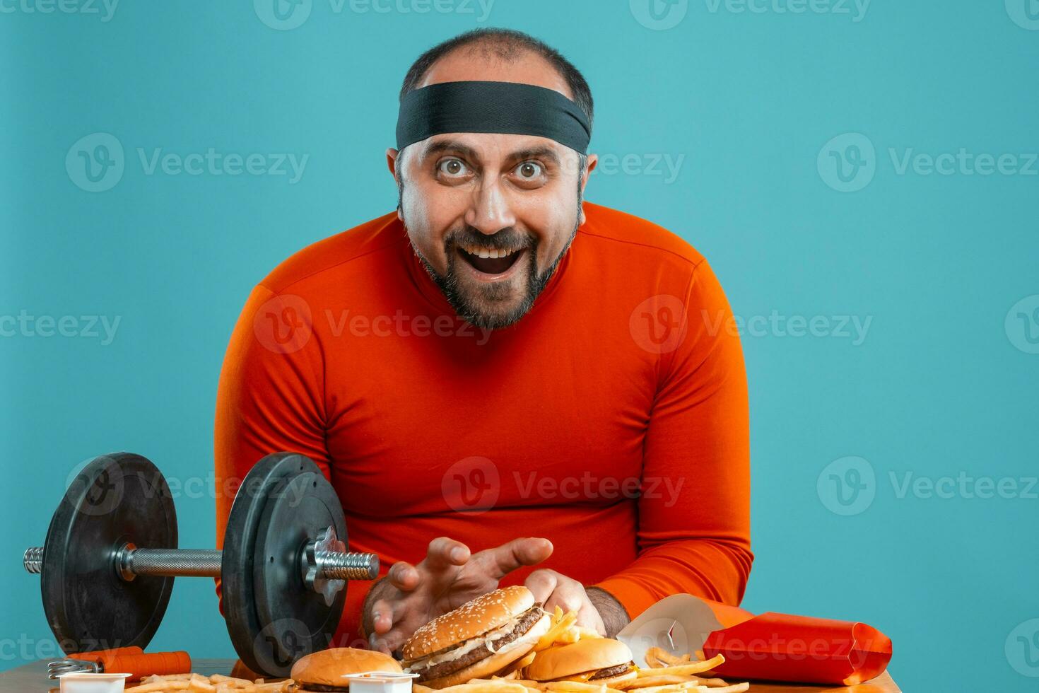 Middle-aged man with beard, dressed in a red turtleneck, headband, posing with burgers and french fries. Blue background. Close-up. Fast food. photo