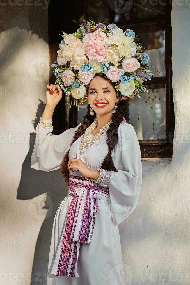 Brunette girl in a white ukrainian authentic national costume and a wreath of flowers is posing against a white hut. Close-up. photo