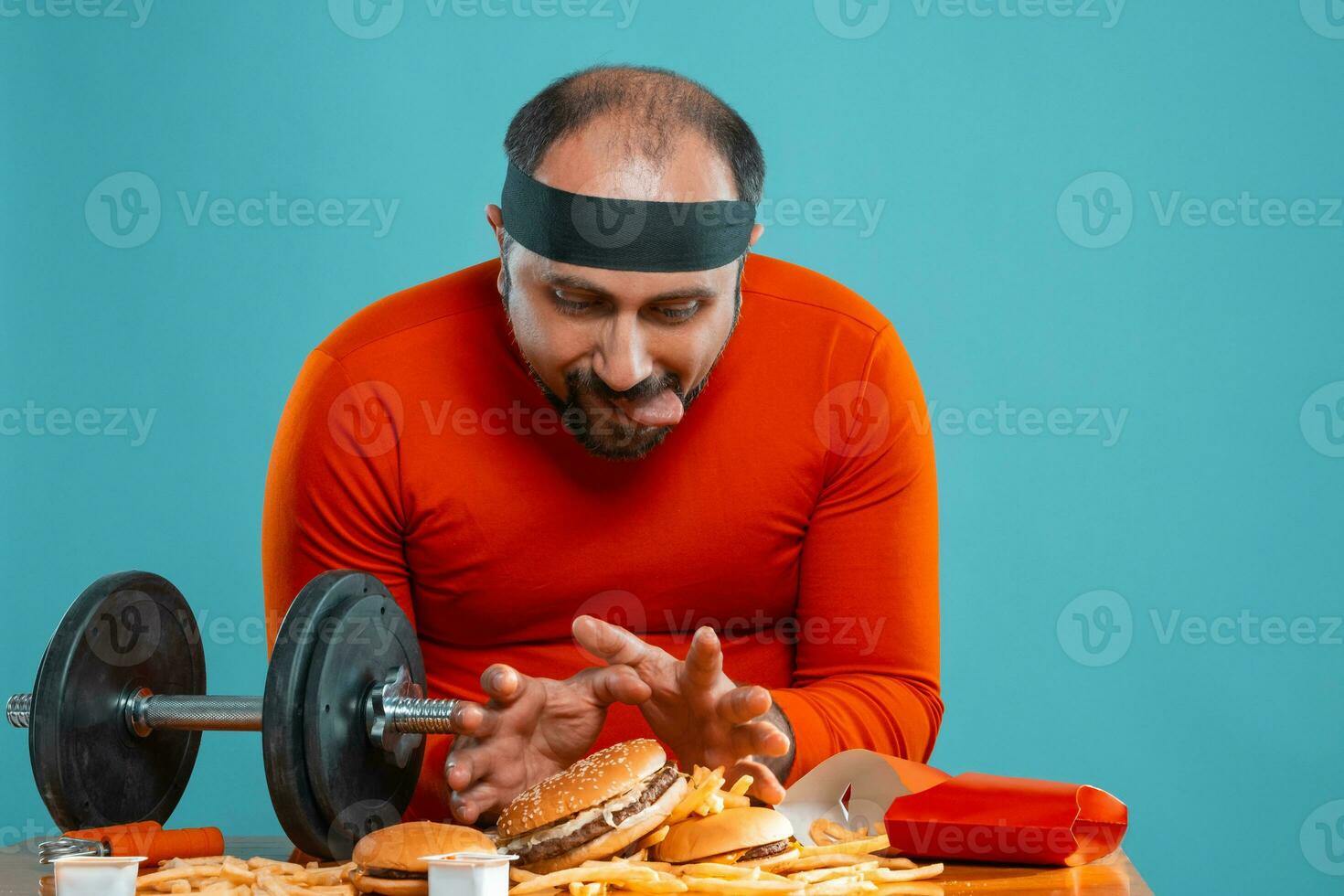 Middle-aged man with beard, dressed in a red turtleneck, headband, posing with burgers and french fries. Blue background. Close-up. Fast food. photo