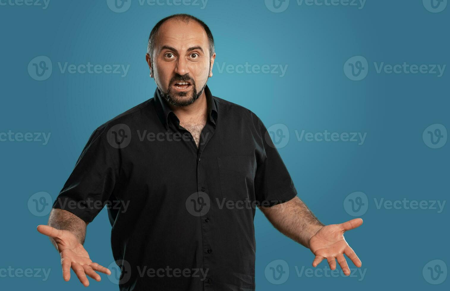 Close-up portrait of a brunet middle-aged man with beard, dressed in a black t-shirt and posing against a blue background. photo