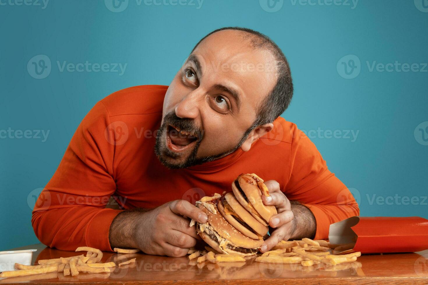 Close-up portrait of a middle-aged man with beard, dressed in a red turtleneck, posing with burgers and french fries. Blue background. Fast food. photo