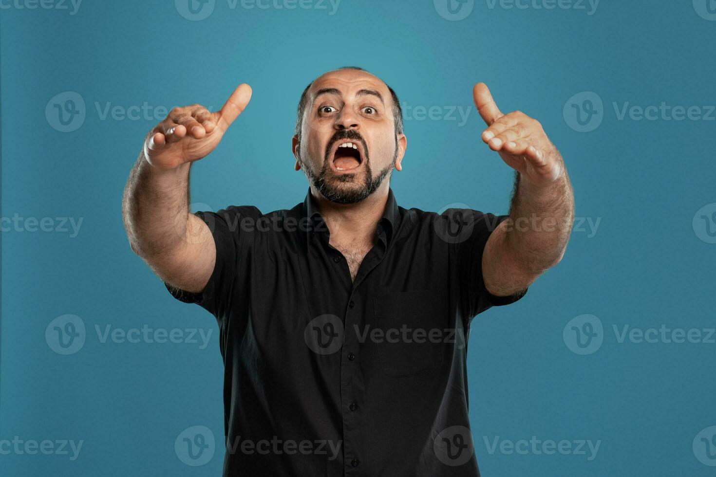 Close-up portrait of a brunet middle-aged man with beard, dressed in a black t-shirt and posing against a blue background. photo