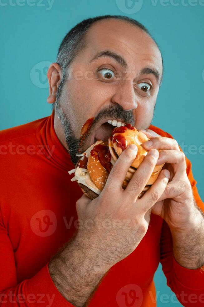 Close-up portrait of a middle-aged man with beard, dressed in a red turtleneck, posing with burgers against a blue background. Fast food. photo